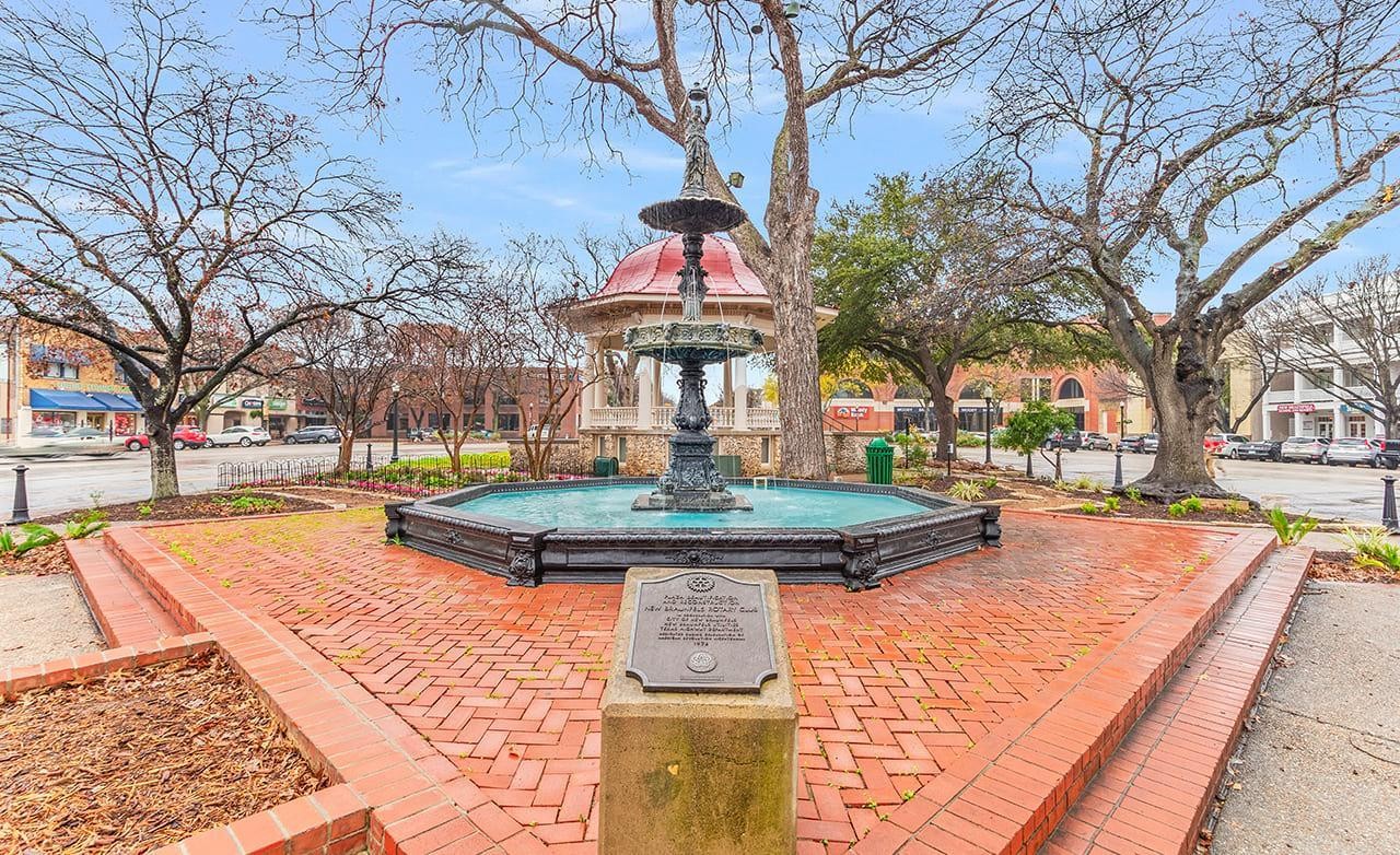 Ornate gazebo and fountain in New Braunfels, Texas, surrounded by brick pathways and lush trees. Urban backdrop.