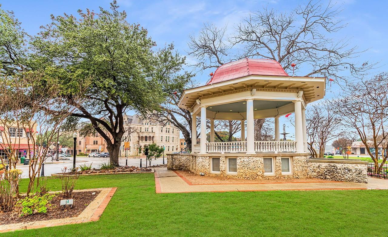 Gazebo with red roof and stone base on lush lawn, trees, courthouse view, Texas. Historic, town square setting.