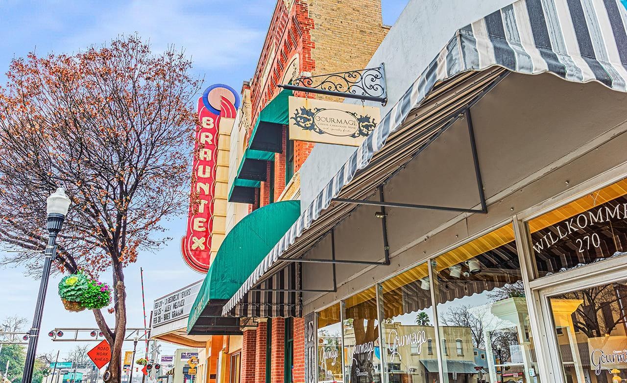 Brauntex Theatre in New Braunfels, Texas with red vintage marquee, adjacent shops, and tree-lined street view.